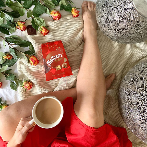 Photo of a woman's hand, holding a cup of tea beside a bag of Maltesers